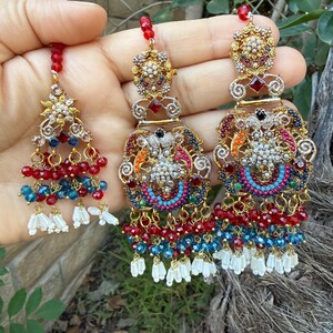 May include: Ornate gold-toned earrings with intricate detailing. The earrings feature red, blue, and white beaded accents, along with dangling white tassels. The design includes floral and geometric patterns, with a central cluster of small, sparkling stones.