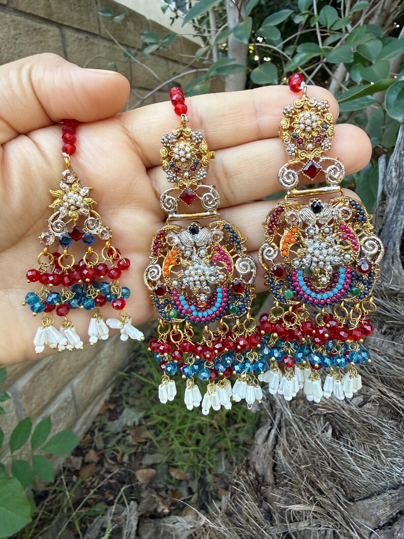 May include: Ornate gold-toned earrings with intricate detailing. The earrings feature red, blue, and white beaded accents, along with dangling white tassels. The design includes floral and geometric patterns, with a central cluster of small, sparkling stones.