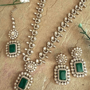 May include: An antique gold-toned jewelry set featuring a necklace, earrings, and a headpiece. The set includes emerald green rectangular stones surrounded by clear, round crystals. The necklace and earrings have a floral design.