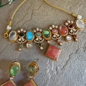 May include: A gold-toned necklace and matching earrings set. The necklace features a series of colorful gemstones, including turquoise, green, and pink, set in ornate gold frames. The earrings have a green oval stone and a pink square stone.