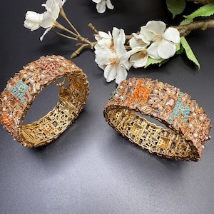 May include: Two gold-colored, ornate bangles with intricate openwork designs. The bangles are adorned with a variety of colorful gemstones, including turquoise, orange, and clear stones. White flowers and branches are in the background.