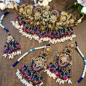 May include: A gold-toned jewelry set featuring a necklace, earrings, and a headpiece. The set is adorned with blue, pink, and white beads, and intricate floral designs. The jewelry is displayed on a wooden surface, with white flowers in the background.