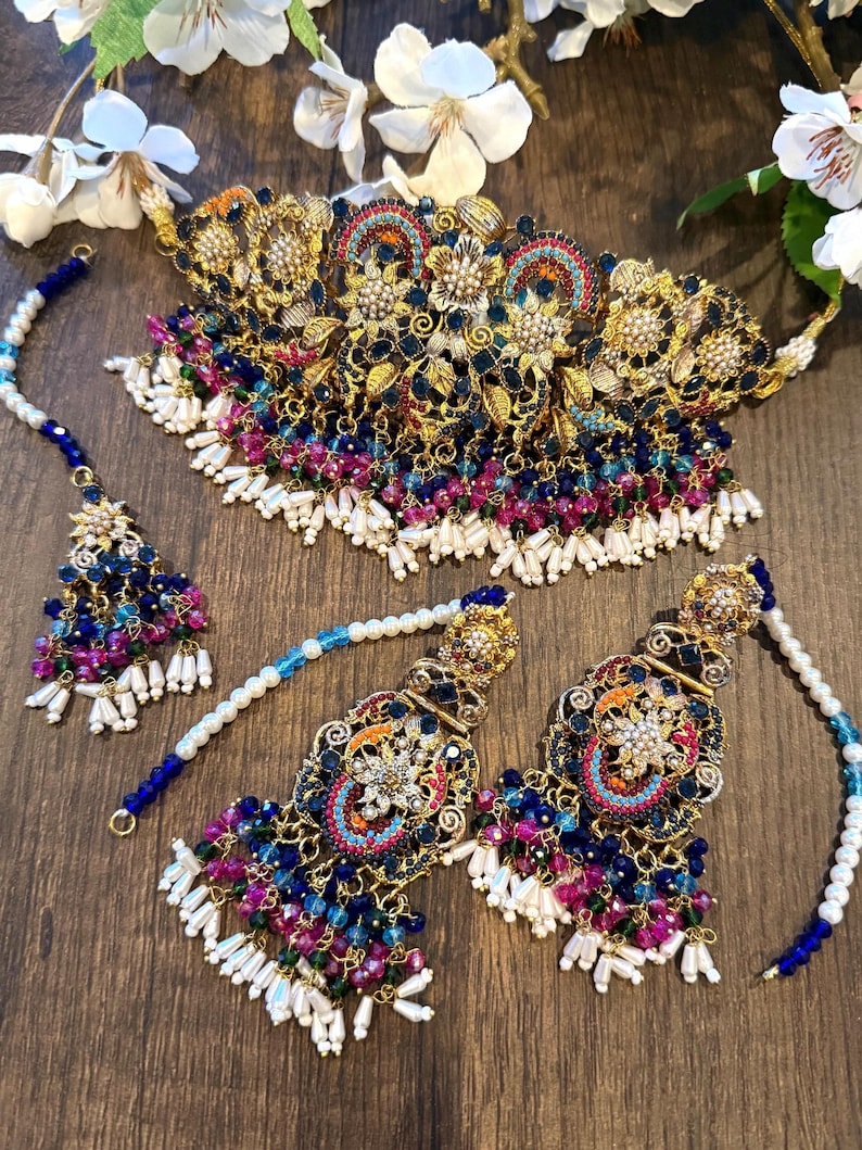 May include: A gold-toned jewelry set featuring a necklace, earrings, and a headpiece. The set is adorned with blue, pink, and white beads, and intricate floral designs. The jewelry is displayed on a wooden surface, with white flowers in the background.
