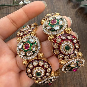 May include: Gold-toned bracelet with ornate, circular designs. Each circle features a central gemstone, surrounded by smaller stones and a border of clear crystals. The bracelet has red and green accents.