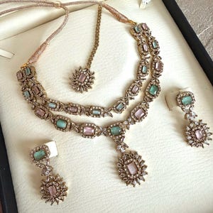 May include: A gold-toned jewelry set featuring a double-strand necklace with alternating rectangular pink and mint green stones, a matching pendant, two drop earrings, and a maang tikka, all accented with small clear crystals in a floral pattern.