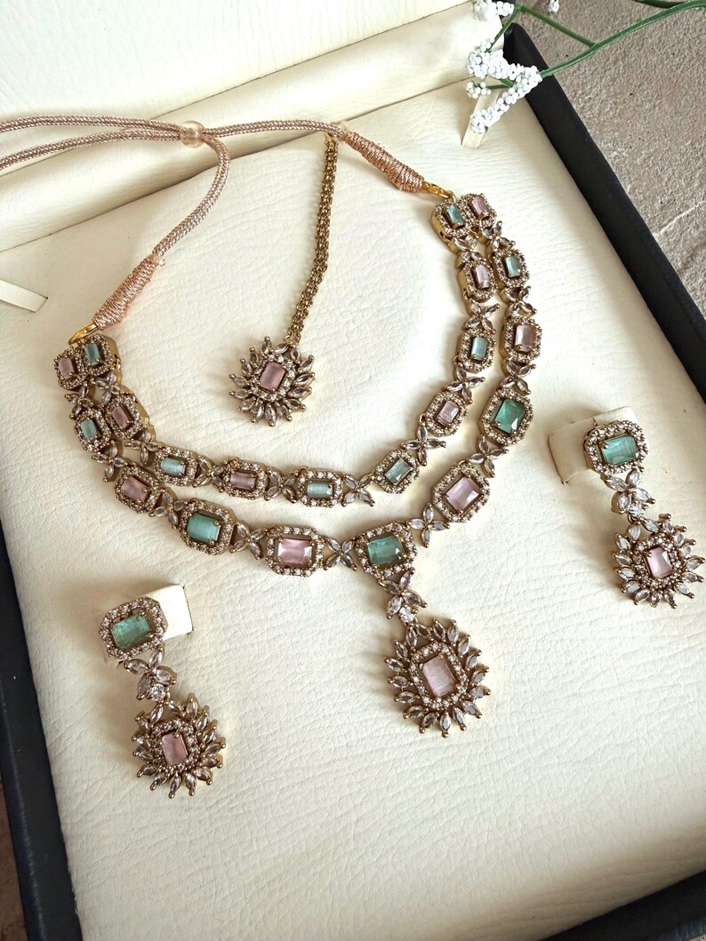 May include: A gold-toned jewelry set featuring a double-strand necklace with alternating rectangular pink and mint green stones, a matching pendant, two drop earrings, and a maang tikka, all accented with small clear crystals in a floral pattern.