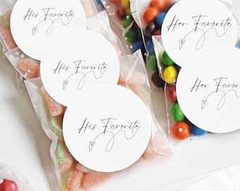 Op de afbeelding: Transparante cellofaan zakjes gevuld met kleurrijke snoepjes, zoals gummy snoepjes en chocolade snoepjes. Witte ronde stickers met de woorden "His Favorite" en "Her Favorite" in zwart schrift zijn op de zakjes bevestigd.