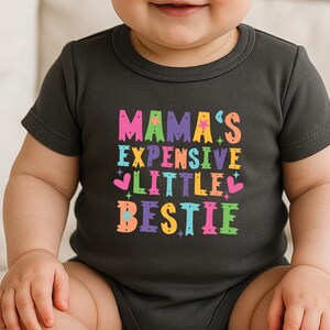 May include: A dark grey heather baby bodysuit featuring the colorful text MAMA'S EXPENSIVE LITTLE BESTIE printed on the front. The text is styled in various bright colors including pink, orange, yellow, green, and blue, accented with small stars and hearts.