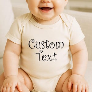 May include: A smiling baby wearing a cream-colored onesie with the words "Custom Text" in black script. The baby also wears a cream-colored bow headband. The baby is sitting on a white couch.