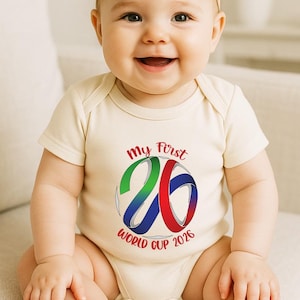 May include: A smiling baby wearing a cream-colored onesie with the text "My First 26 World Cup 2026" in red, green, blue, and white. The baby has a cream-colored bow headband.