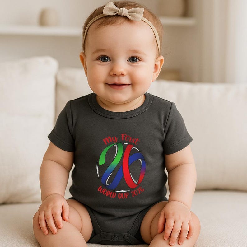 May include: A dark gray baby bodysuit with the text "My First World Cup 2026" in red, green, and blue. The design features a stylized number 26. The baby is wearing a beige bow headband.
