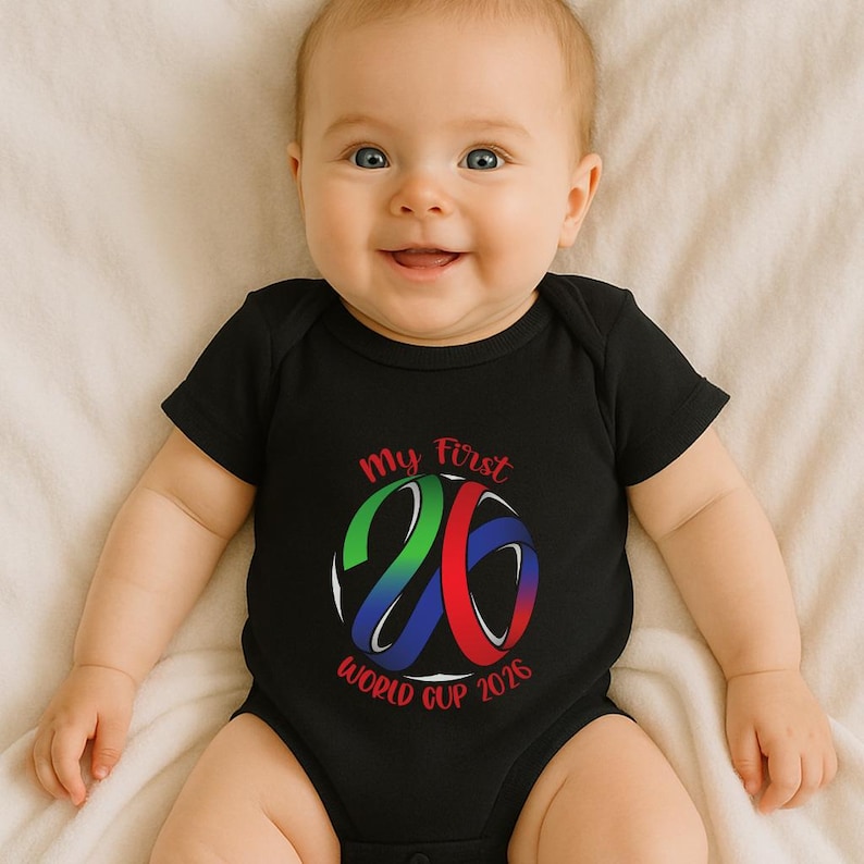 May include: A black baby bodysuit with the text "My First 26 World Cup 2026" in red, green, and blue. The design features a stylized number 26 with a soccer ball theme. The baby is smiling and lying on a white surface.