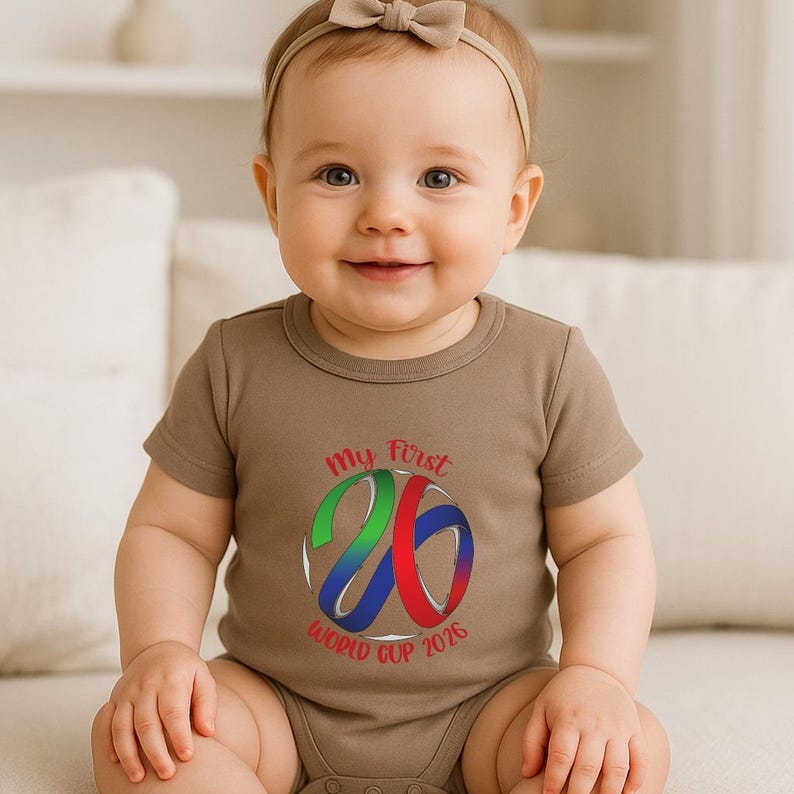 May include: A smiling baby wearing a brown onesie with the text "My First 26 World Cup 2026" in a colorful design. The baby has a beige bow headband.