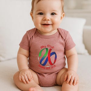 May include: A baby wearing a dusty rose onesie with the text "My First World Cup 2026" in green, blue, and red. The baby is smiling and sitting on a white surface.