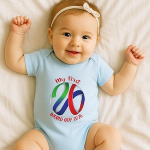 May include: A light blue baby onesie with the text "My First 26 World Cup 2026" in red, green, and blue. The onesie features a colorful graphic of the number 26. The baby is wearing a white headband with a bow.