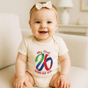 Puede incluir: Un beb&eacute; sonriente con un body color crema con el texto "My First 26 World Cup 2026" en rojo, verde, azul y blanco. El beb&eacute; lleva una diadema con lazo color crema.