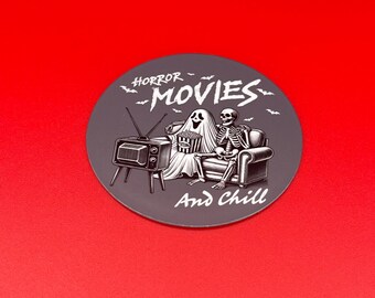 Peut inclure: Un dessous-de-verre rond gris avec une illustration en noir et blanc d'un squelette et d'un fantôme regardant la télévision sur un canapé. Le texte "HORROR MOVIES And Chill" est imprimé sur le dessous-de-verre.