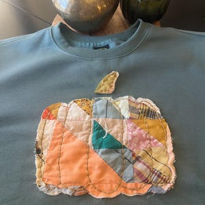以下が含まれることがあります： パッチワークの南瓜のアップリケが付いたターコイズブルーのスウェットシャツ。南瓜はピンク、オレンジ、ブルー、グリーン、イエローの様々な生地で作られています。