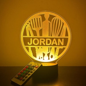 May include: A white personalised night light with a circular design featuring kitchen utensils and the name "JORDAN". The light is on and illuminating the design. The light comes with a remote control.