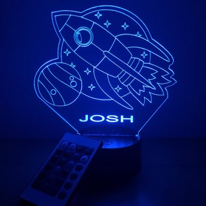 May include: Blue LED night light with a rocket, planet, and stars design. The name "JOSH" is displayed below the design. The night light comes with a remote control.
