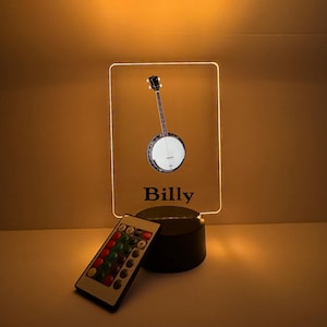 May include: A rectangular LED lamp featuring a banjo illustration and the name "Billy" in black font. The lamp includes a remote control with multiple color options. The base is black.
