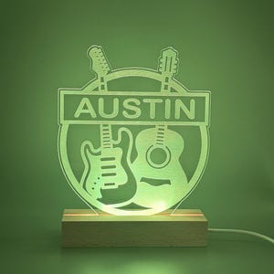 May include: A light-up acrylic sign featuring the name AUSTIN in a rectangular banner, with silhouettes of an electric guitar and an acoustic guitar behind it. The design is mounted on a rectangular wooden base and emits a soft green glow.