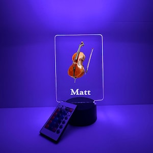Könnte beinhalten: LED-Nachtlicht mit Cello-Motiv und dem Namen "Matt" auf einer transparenten Acrylplatte. Die Leuchte hat einen schwarzen Sockel und eine Fernbedienung zur Farbeinstellung. Der Hintergrund ist ein leuchtendes Lila.