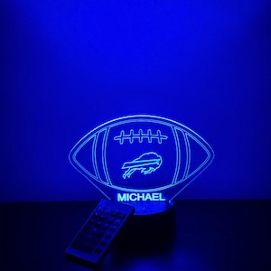 Pode incluir: Lâmpada LED azul em forma de bola de futebol, com o logotipo dos Buffalo Bills e o nome "MICHAEL". Um controle remoto está colocado em frente à lâmpada. A lâmpada emite um brilho azul brilhante contra um fundo escuro.