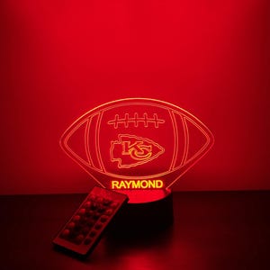 May include: A red illuminated football-shaped lamp with the text "RAYMOND" at the base. The lamp features the Kansas City Chiefs logo. A remote control is placed to the left of the lamp.
