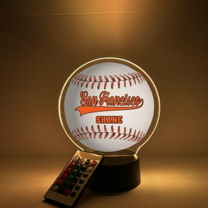 Pode incluir: Lâmpada LED com tema de beisebol com o texto "San Francisco" e "Shane" em laranja. A lâmpada tem um design de bola de beisebol branca com costuras vermelhas e um controle remoto.