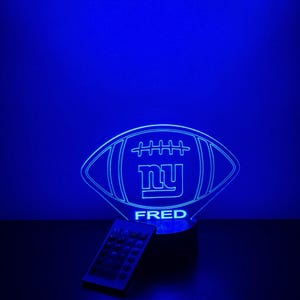 May include: A blue LED lamp shaped like a football, with the letters "NY" inside and the name "FRED" below. A remote control is placed to the left of the lamp. The lamp emits a bright blue glow.