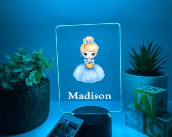 Könnte beinhalten: Personalisiertes LED-Nachtlicht mit einer Cartoon-Prinzessin, die einen Kürbis hält, mit dem Namen "Madison" darunter. Das Licht hat einen blauen Schein, eine Fernbedienung und steht auf einem schwarzen Sockel. Dekorative Pflanzen und ABC-Blöcke sind im Hintergrund.