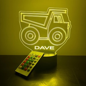 Puede incluir: Una luz nocturna LED amarilla con forma de camión volquete con el nombre "DAVE" debajo. La luz está sobre una base negra y viene con un mando a distancia.