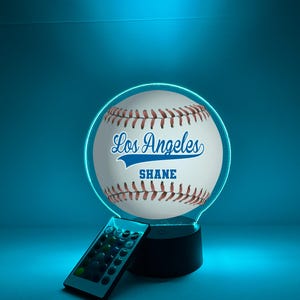 May include: An illuminated baseball-themed lamp. The design features a white baseball with red stitching, the text "Los Angeles" and "SHANE" in blue. It has a black base and a remote control. The background is a blue gradient.
