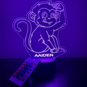 Puede incluir: Una luz nocturna morada con forma de mono con el nombre "Aiden" impreso debajo. La luz nocturna se controla con un mando a distancia con varios botones de colores.