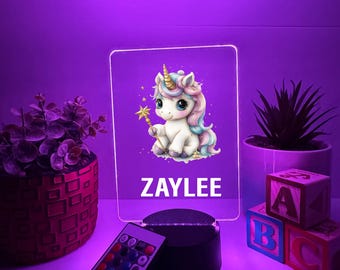 May include: A white unicorn with a rainbow mane and a gold horn holding a wand. The unicorn is on a light blue background with stars. The name "ZAYLEE" is written below the unicorn. The image is on a clear acrylic panel with a black base and a remote control.
