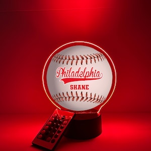 Pode incluir: Lâmpada LED em forma de bola de beisebol com a palavra "Philadelphia" e o nome "Shane" em escrita vermelha. A lâmpada tem um design de beisebol branco com costuras vermelhas e assenta numa base preta. Um controlo remoto está em primeiro plano.