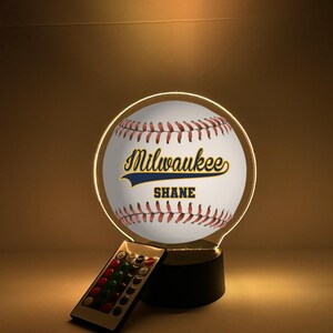 Pode incluir: Luz noturna com tema de beisebol com o texto "Milwaukee" e "Shane" na bola. A luz tem um design circular com uma borda brilhante e um controle remoto.