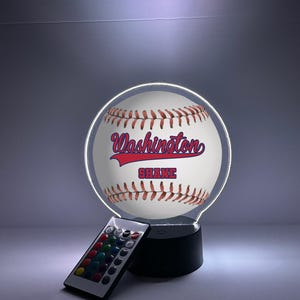 Pode incluir: Luz noturna com tema de beisebol com os textos "Washington" e "Shane" em vermelho e azul. A luz tem um design de bola de beisebol com costuras vermelhas e vem com um controle remoto para ajustes de cor. A base é preta.