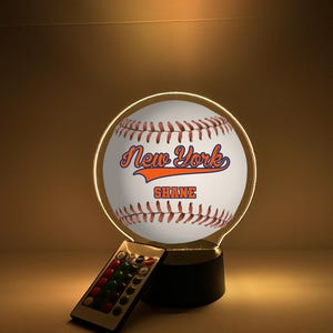 Pode incluir: Lâmpada LED em forma de bola de beisebol com os textos "New York" e "Shane" em laranja e azul. O design inclui costuras marrons e um controle remoto. A base é preta.