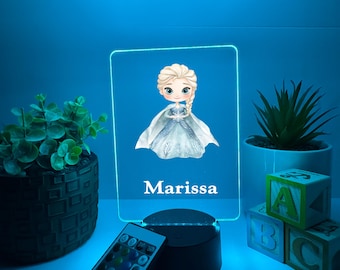 Könnte beinhalten: Rechteckiges LED-Nachtlicht mit einem Cartoon-Bild einer Prinzessin in einem blauen Kleid und dem Namen "Marissa" darunter. Das Licht steht auf einem schwarzen Sockel, mit einer Fernbedienung daneben. Dekorative Pflanzen und Buchstabenblöcke im Hintergrund.