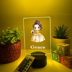 Op de afbeelding: Gepersonaliseerde LED-nachtlamp met een cartoonillustratie van een prinses die een roos vasthoudt. De naam "Grace" staat onder de afbeelding. Inclusief afstandsbediening en een zwarte voet. Decoratieve planten en ABC-blokken staan op de achtergrond.