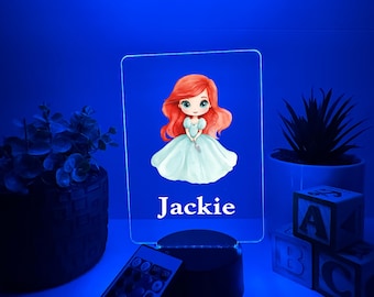 Könnte beinhalten: Rechteckiges LED-Nachtlicht mit einer Cartoon-Illustration einer rothaarigen Prinzessin in einem blauen Kleid. Der Name "Jackie" ist weiß gedruckt. Inklusive Fernbedienung und schwarzem Sockel. Blaues Licht.
