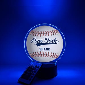 Pode incluir: Lâmpada LED com tema de beisebol com design de bola de beisebol branca, costuras vermelhas e o texto "New York" e "SHANE" em azul marinho. A lâmpada tem um contorno de néon azul e vem com um controle remoto.
