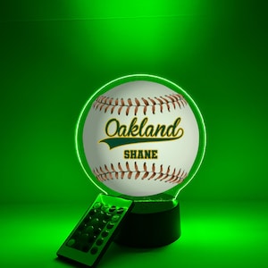 Pode incluir: Uma luz noturna com tema de beisebol com um contorno de néon verde. A bola de beisebol tem costuras marrons e o texto "Oakland" e "SHANE" em verde e branco. Um controle remoto está na frente da luz.