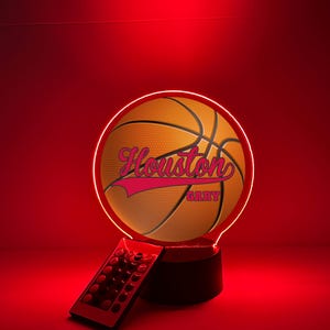 Puede incluir: Lámpara LED con temática de baloncesto con la palabra "Houston" en escritura rosa en la superficie de la pelota. La lámpara tiene un brillo rojo e incluye un mando a distancia. La base es negra.