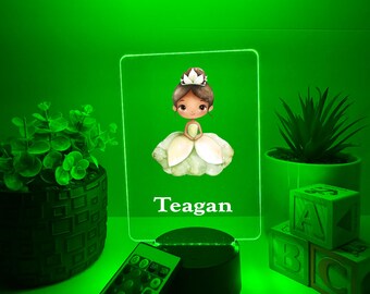 Könnte beinhalten: Grün beleuchtetes Acryl-Nachtlicht mit einer Cartoon-Ballerina und dem Namen "Teagan". Das Licht steht auf einem schwarzen Sockel, mit einer Fernbedienung. Dekorative Pflanzen und ABC-Blöcke befinden sich im Hintergrund.