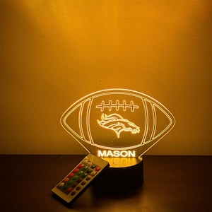 May include: A glowing football-shaped LED night light with the name "MASON" and a team logo. The light is controlled by a remote with multiple color options. The light is on a dark surface.