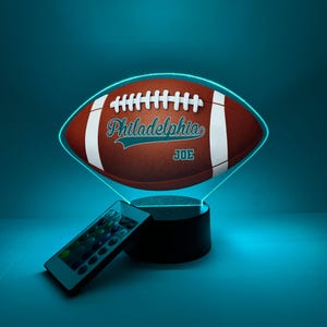 May include: A brown football-shaped LED lamp with the text "Philadelphia" and "JOE" in teal. The lamp has a white outline and sits on a black base, with a remote control nearby. The background is a teal color.