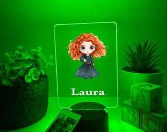 Könnte beinhalten: Grün beleuchtetes LED-Nachtlicht mit einer Cartoon-Illustration eines Charakters mit roten Locken und dem Namen "Laura". Das Licht wird von einer Fernbedienung begleitet und steht auf einem schwarzen Sockel. Dekorative Pflanzen und ABC-Blöcke sind ebenfalls sichtbar.